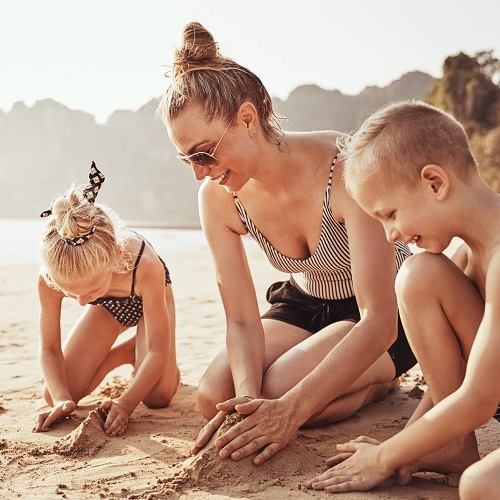Smiling Mom and her two adorable children playing in the sand together during summer vacation at the beach